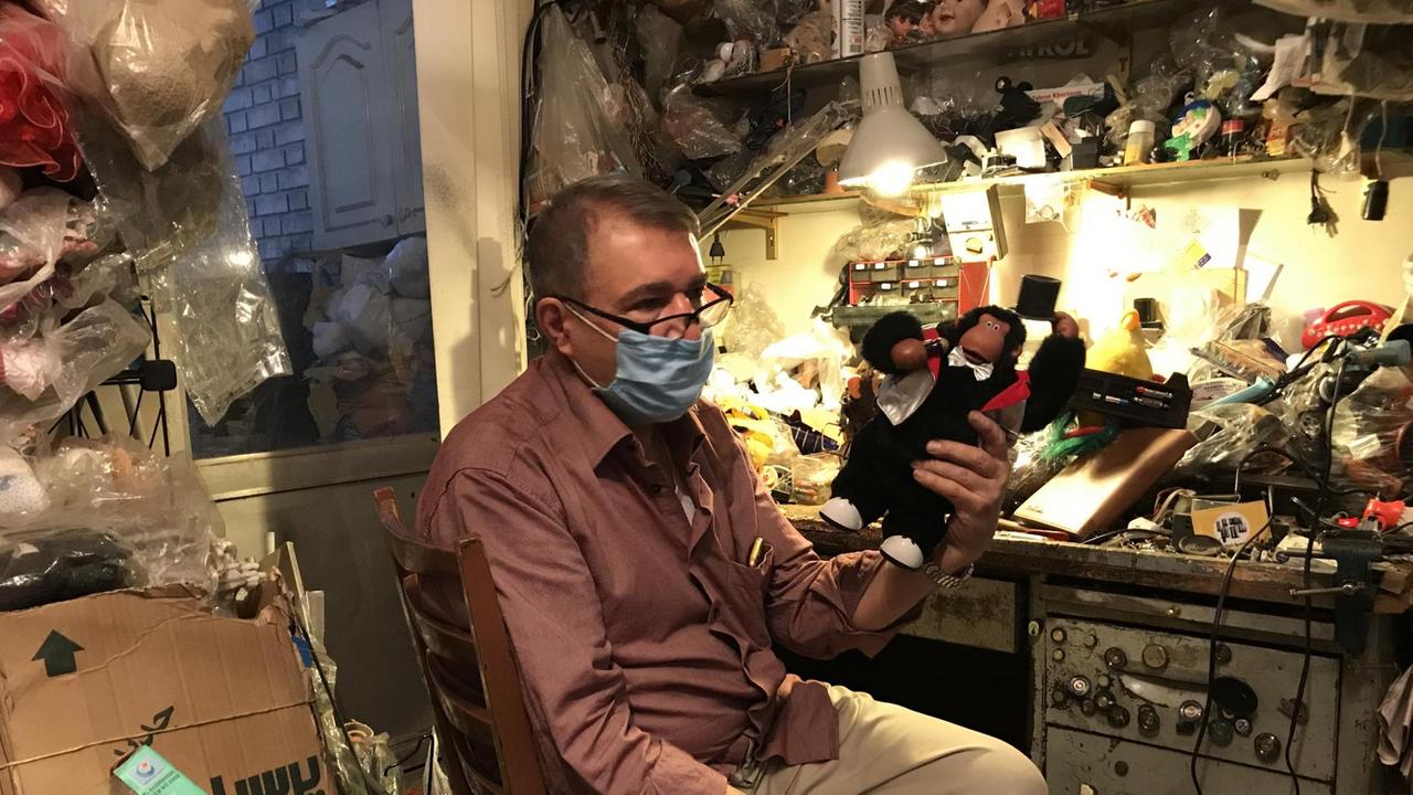Ein älterer Herr mit Mundschutz sitzt in einem kleinen Atelier umgeben von jeder Menge Spielzeug und hält einen Spielzeugaffen in der Hand.