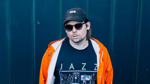 Ein junger Mann mit Kappe, Sonnenbrille und orangener Regenjacke steht vor einer dunkelblauen Wand. Er trägt ein T-Shirt mit der Aufschrift "Jazz".