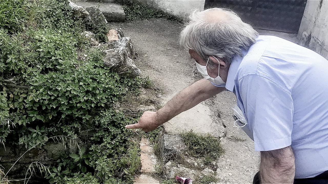 Ein älterer Herr in weißem Hemd mit Corona-Schutzmaske zeigt auf ein halb von Pflanzen verdecktes Loch hinter einem Gebäude.