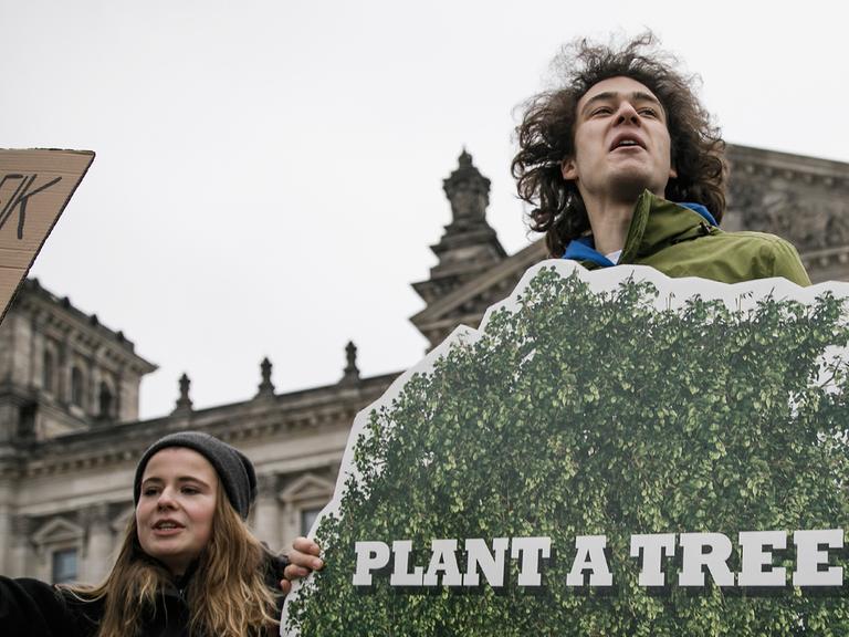 Ein junger Mann steht vor dem Bundestag und hält ein Plakat hoch, auf dem steht: "Plant a tree"