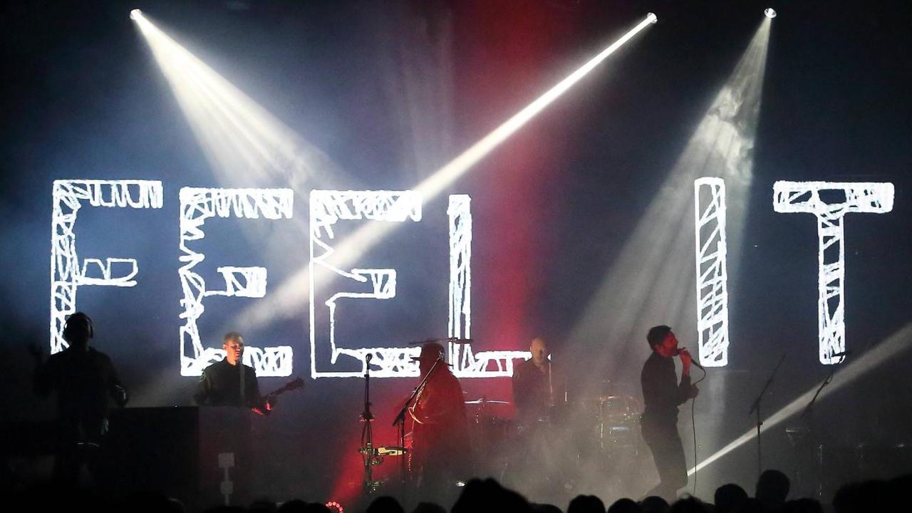 Fünf Männer stehen auf einer Bühne und spielen Musik. Auf der Bühne sind die Worte "Feel it" zu lesen.