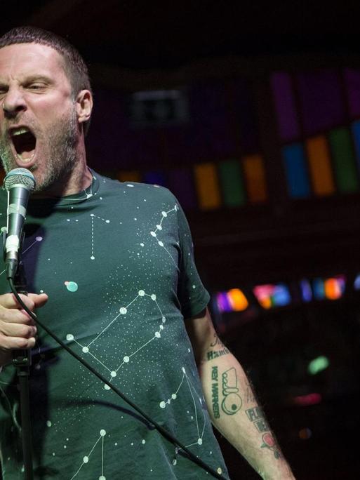 Ein Mann mit Dreitagebart schreit auf einer Bühne in ein Mikrofon. Er trägt ein bedrucktes dunkelgraues T-Shirt. Auf den Unterarmen sind viele verschiedene Tattoos zu erkennen.