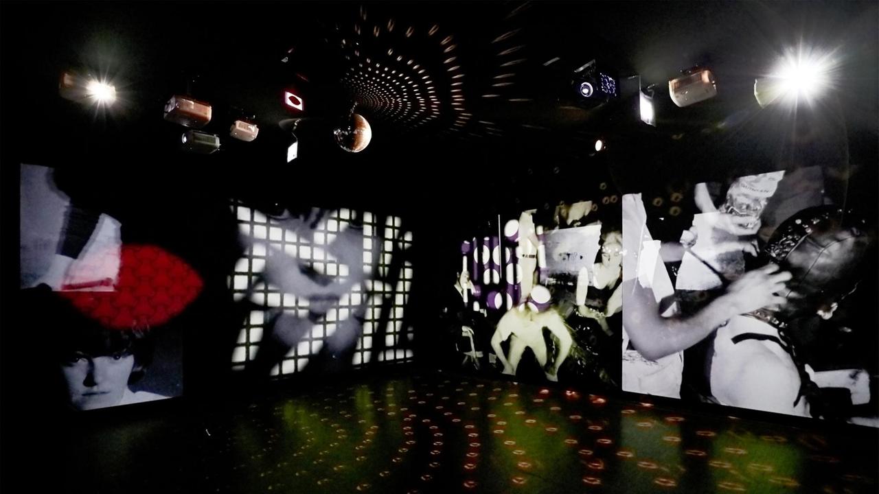 
Installationsansicht der Ausstellung "Andy Warhol Now" im Museum Ludwig, Köln 2021. In dem in schwarz gehaltenen, dunklen Raum sind verschiedene Videoinstallationen an den Wänden zu sehen. 
