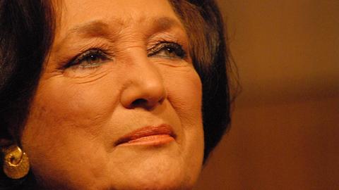 Nahaufnahme einer älteren Dame mit dunkelbraunem, kurzen Haar und braunen Augen blickt nachdenklich zur Seite. Sie trägt goldene Ohrringe, blauen Kajal und roséfarbenen Lippenstift.