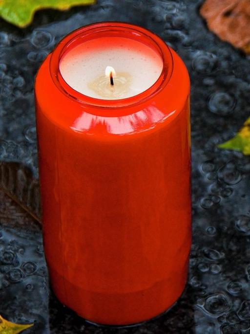 Ein rotes Grablicht steht auf einer grauen Schieferplatte und ist von herbstlich gefärbten Blättern umgeben.