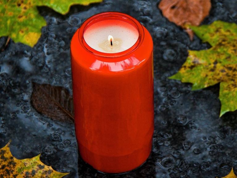 Ein rotes Grablicht steht auf einer grauen Schieferplatte und ist von herbstlich gefärbten Blättern umgeben.