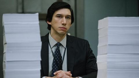 Adam Driver sitzt am Schreibtisch hinter zwei großen Papierstapeln