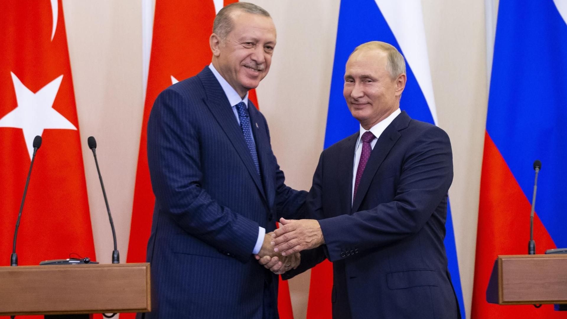 Russlands Präsident Putin (rechts) und der türkische Staatschef Erdogan geben sich die Hand und lächeln in die Kamera. Hinter ihnen sind die Flaggen beider Länder zu sehen.