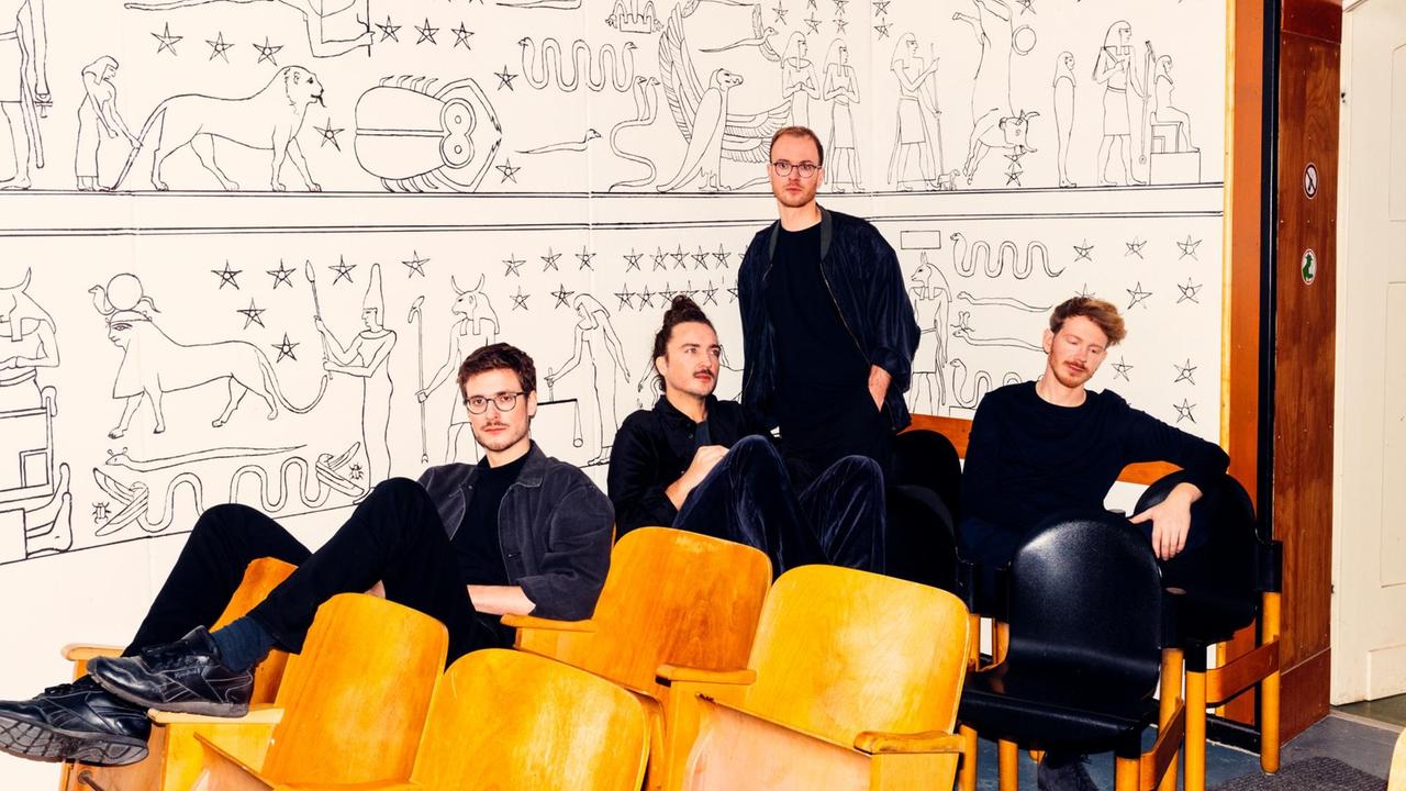 Vier junge Musiker lümmeln in hölzernen Kinostühlen vor weißer Wand mit ägyptischen Motiven 
