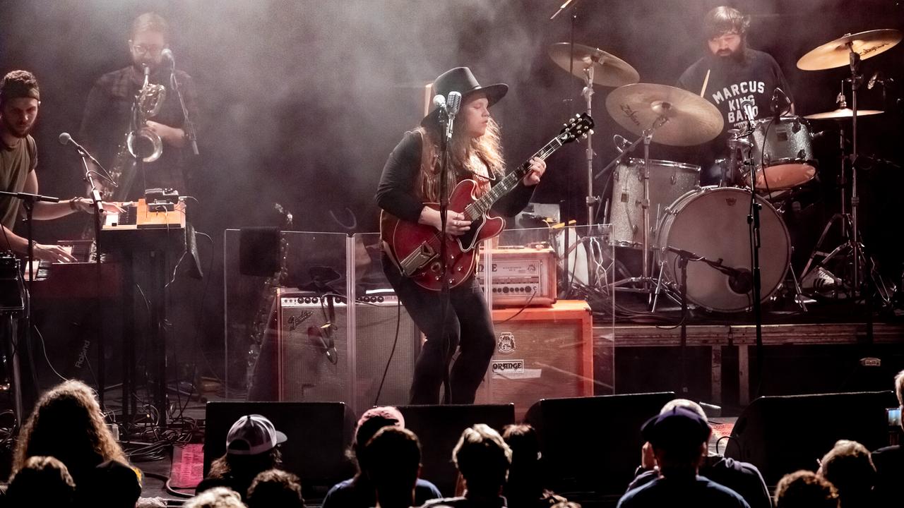 Vier Musiker stehen auf einer Bühne, der Gitarrist und Sänger in der Mitte mit roter Gitarre vor seinem Verstärker und spielt mit geschlossenen Augen.