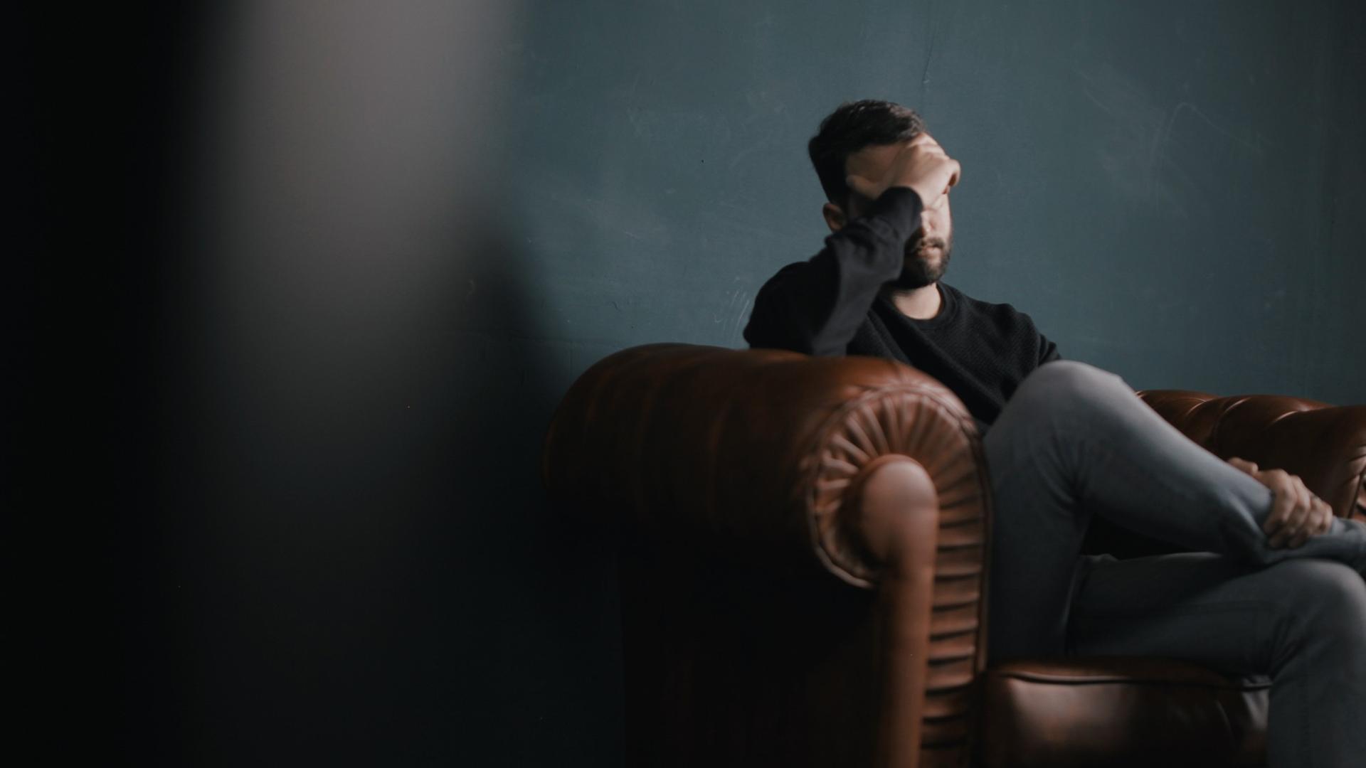  Ein Mann sitzt traurig auf einem Sofa und hält die Hand vor sein Gesicht.