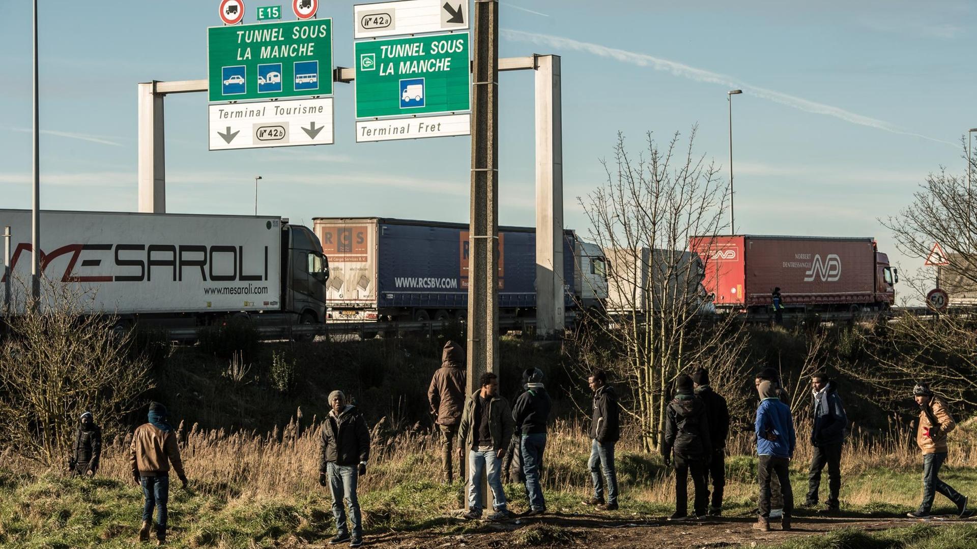 Flüchtlinge stehen am Rande einer Autobahn, auf der Lkws fahren. Über der Straße weist ein Schild Richtung Eurotunnel.