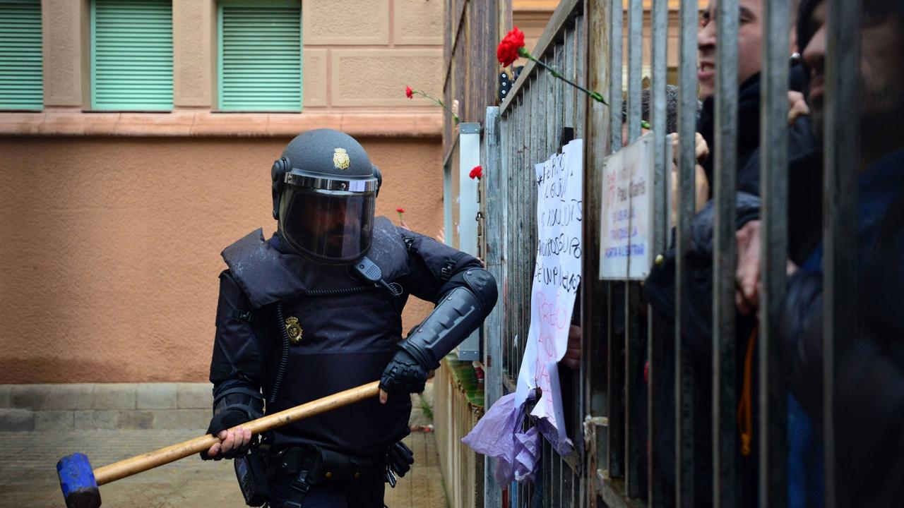 Spanische Polizisten versuchen in die Pau Claris Schule in Barcelona einzudringen, in der über das katalonische Referendum abgestimmt wird. Man sieht einen Polizisten mit schussicherer Weste und mit geschlossenem Helm, der mit einem riesigen Hammer vor dem vergitterten Eingang der Schule steht und offenbar zum Schlag ausholt.