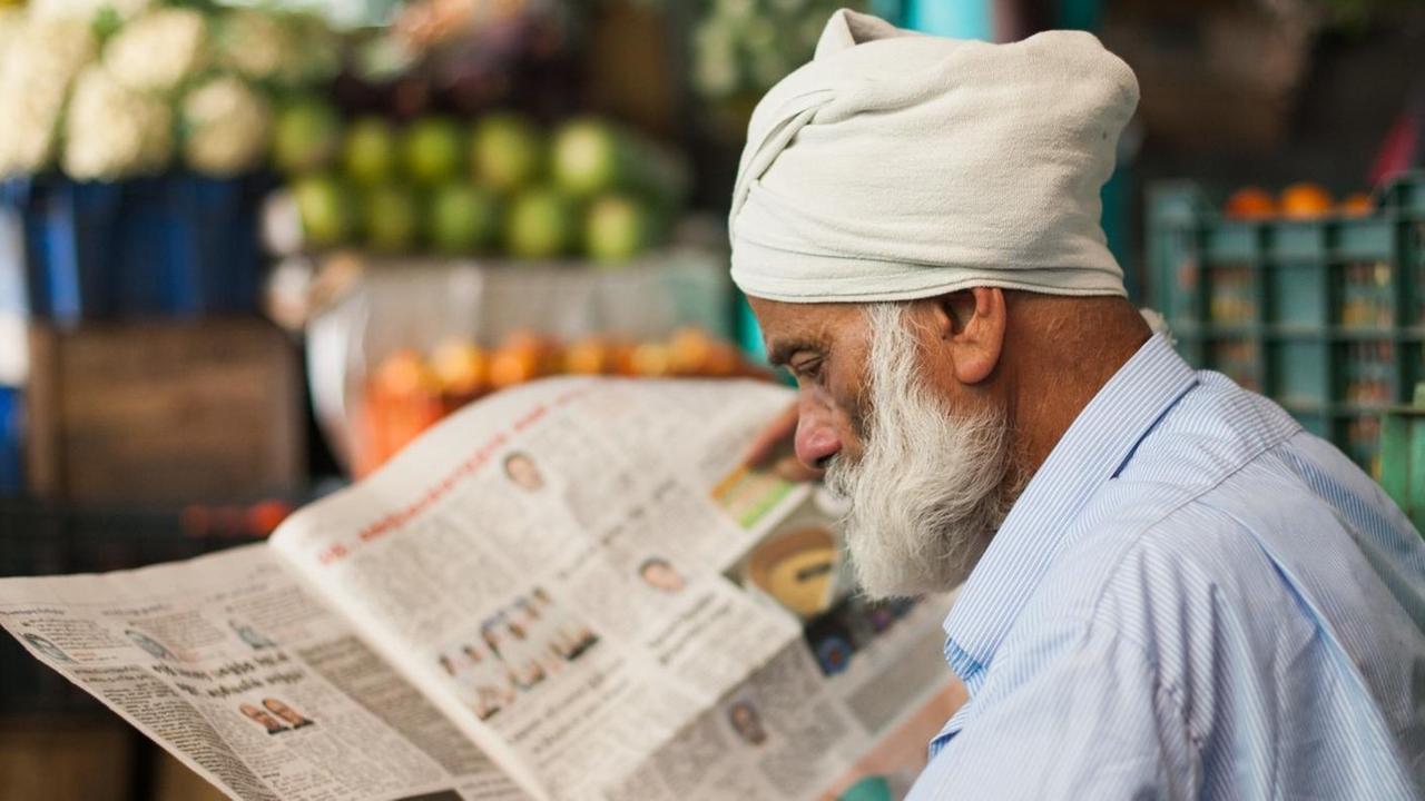Der alte Mann mit Zeitung trägt einen weißen Bart und einen weißen Turban. Er sitzt vor einem Marktstand mit Obst.