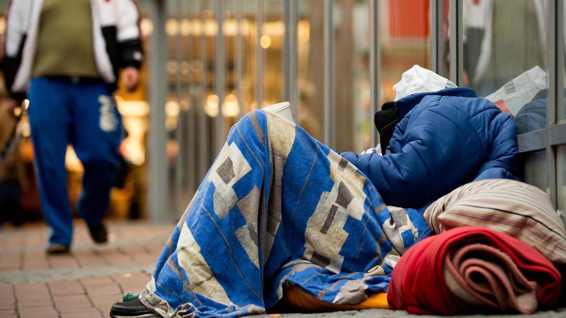 Ein Obdachloser schläft in einer Fußgängerzone in Dortmund, eingepackt in Decken und zwischen Plastiktüten auf dem Boden, während ein Mann vorbei läuft.