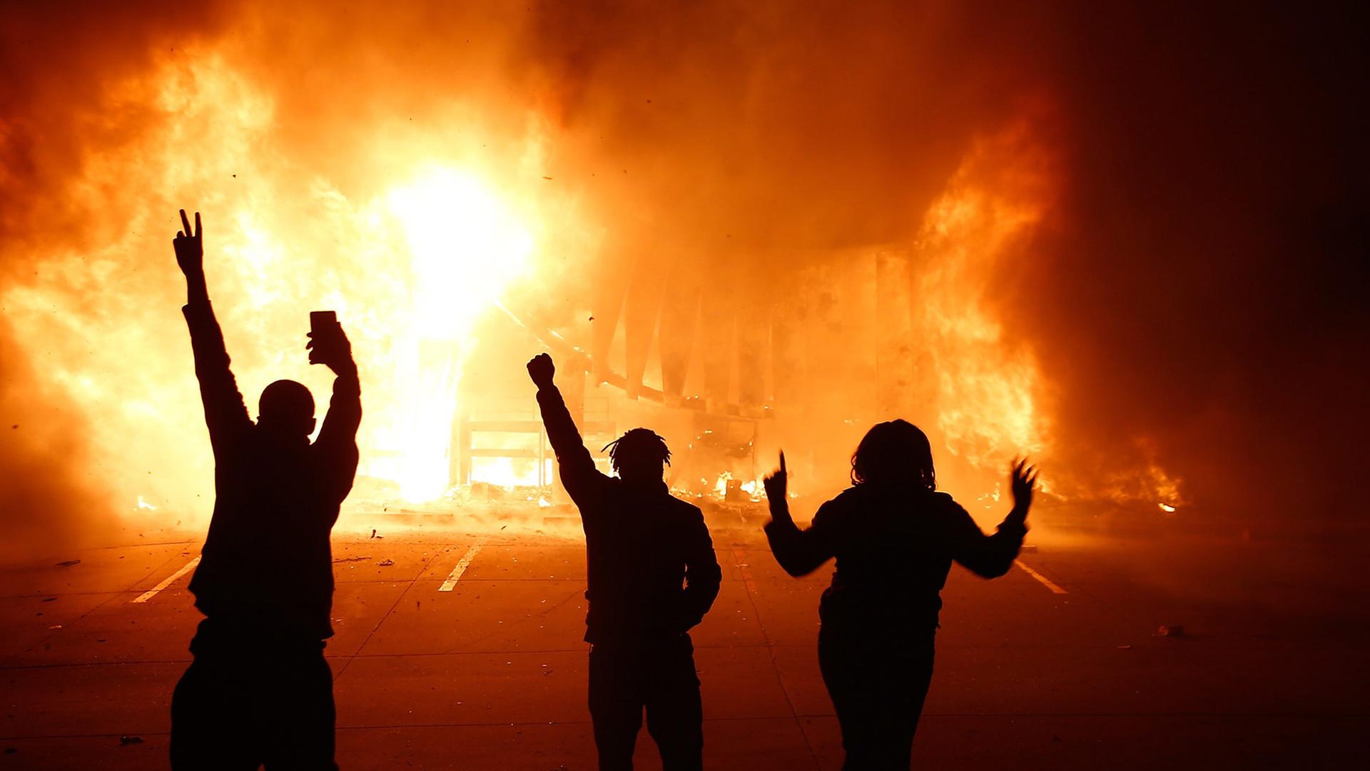Drei Protestierende auf der Straße, im Hintergrund brennt es.