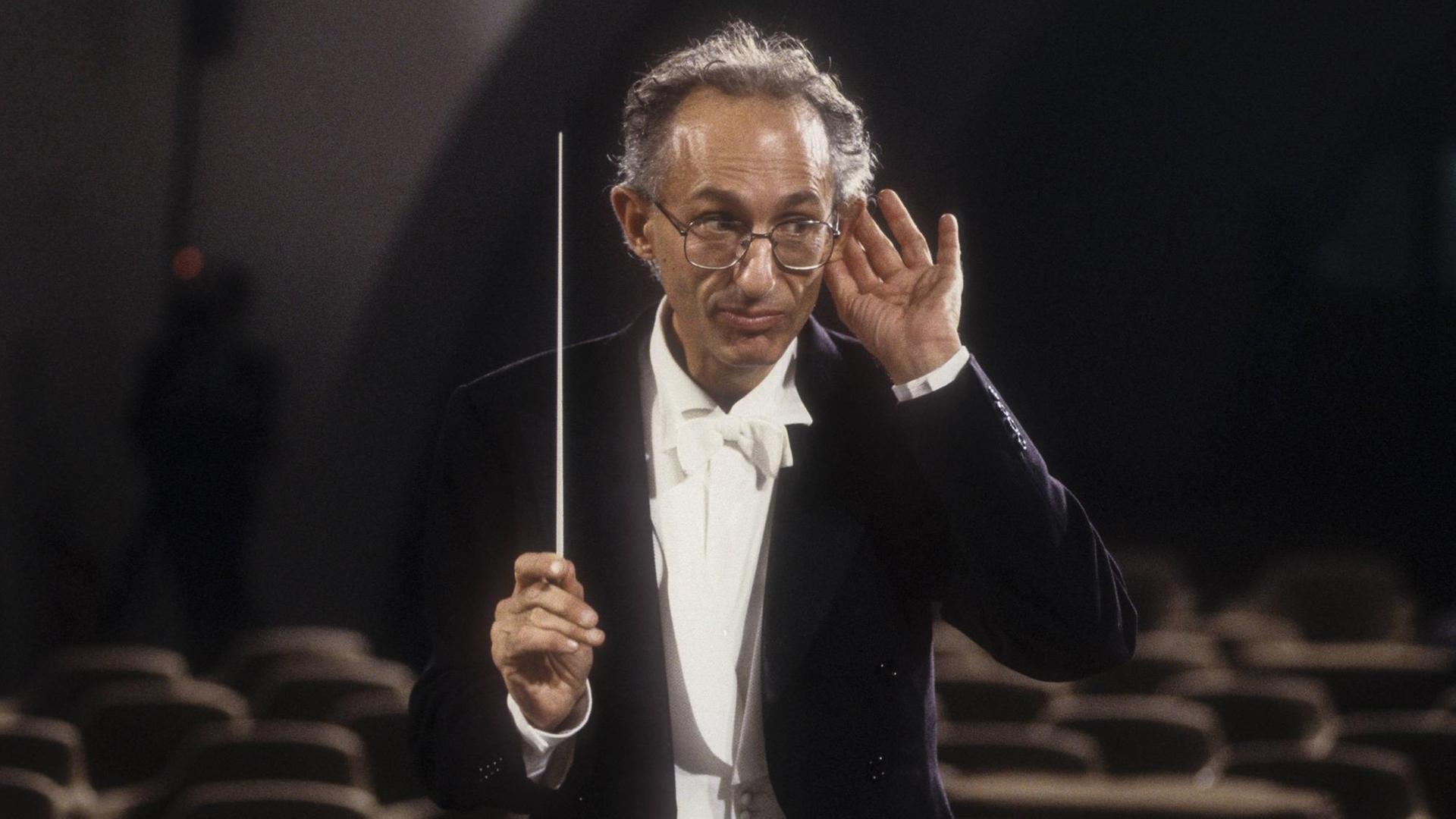 Der italienische Dirigent Claudio Scimone (mit Brille) im Konzertfrack mit weißer Fliege dirigiert mit einem Taktstock und macht eine Horch-Geste mit der linken Hand