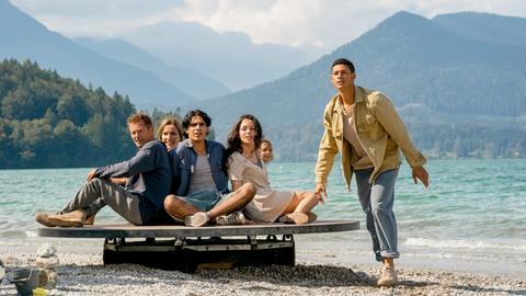 Szenenbild aus "Die Rettung der uns bekannten Welt". Eine Personengruppe ist mit einem Floß am Ufer gestrandet. Im Hintergrund ist ein Bergpanorama mit See zu sehen.