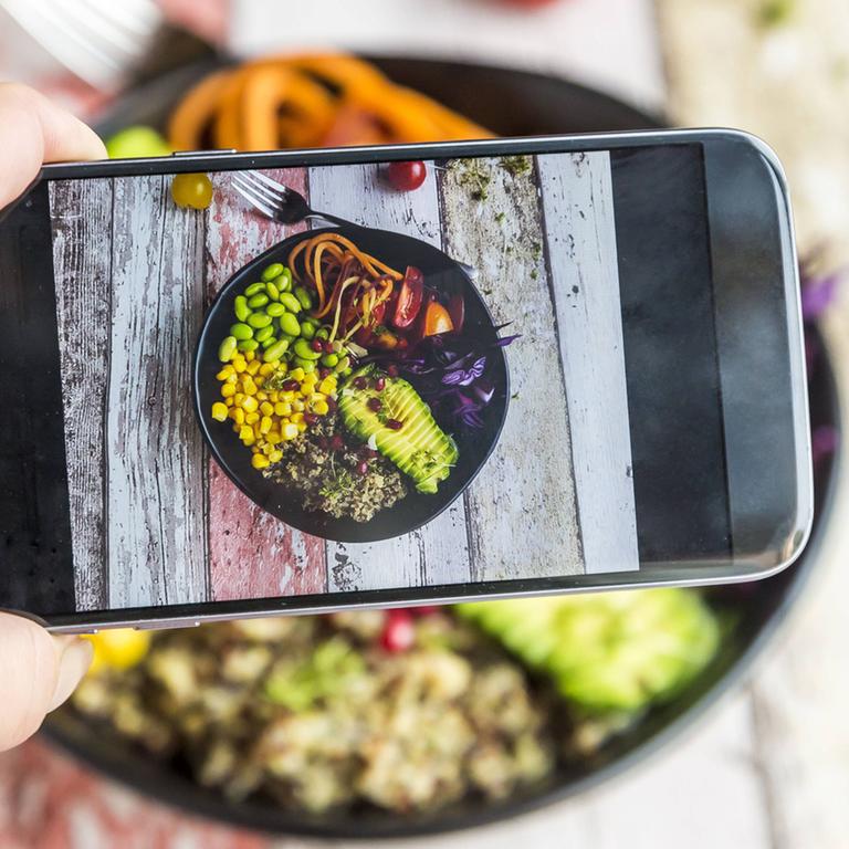 Eine Frau fotografiert ihre vegane Lunch bowl mit ihrem Smartphone.