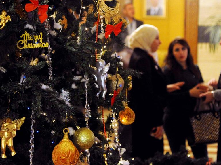 Ein Weihnachtsbaum im syrischen Damaskus. Im Hintergrund sieht man drei Frauen, die sich unterhalten. Zwei von ihnen tragen weiße Kopftücher.