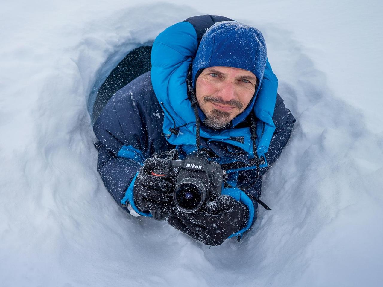 Markus Mauthe in blauer Mütze, Winterjacke, Handschuhen, Fotokamera schaut aus einem Eisloch heraus nach oben in die KAmera.  