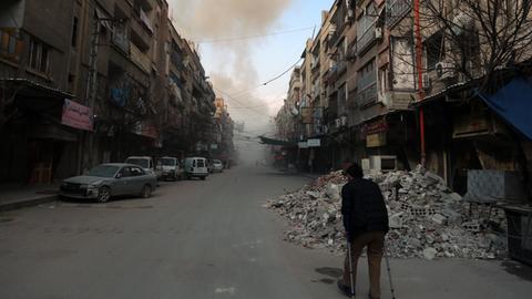 Ost-Ghouta: Ein Mann läuft in der Stadt Douma eine mit Trümmern übersäte Straße entlang, im Hintergrund sind Rauchwollken zu sehen.