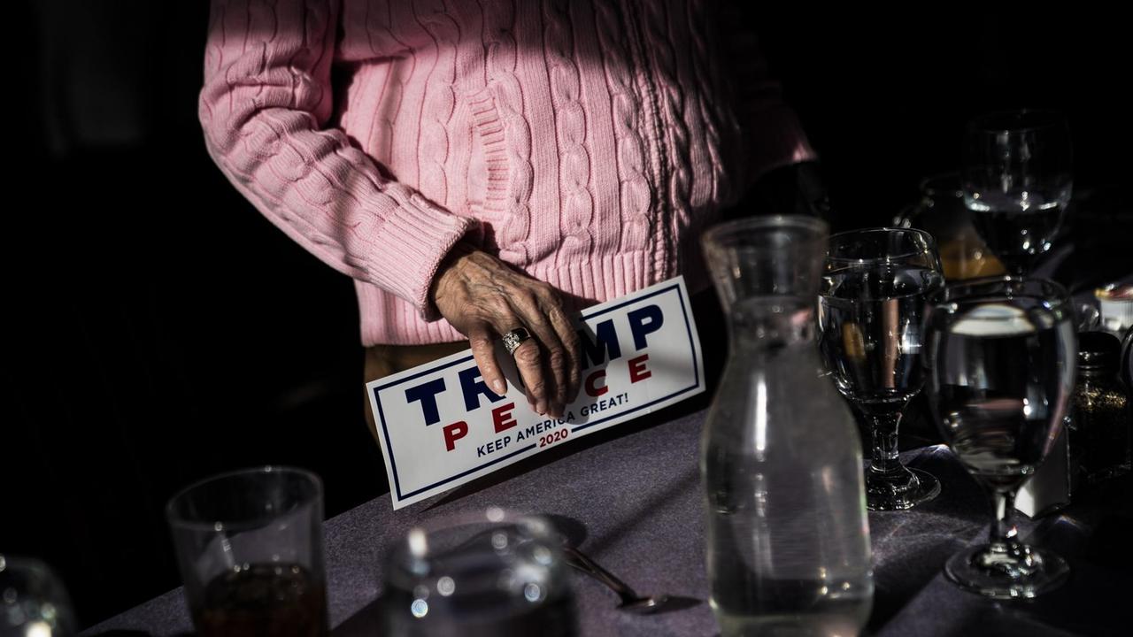 Die Hand einer älteren Frau in einem rosafarbenen Pullower. Auf dem Schild ist die Aufschrift "Trump Pence Keep America Great 2020" zu sehen.