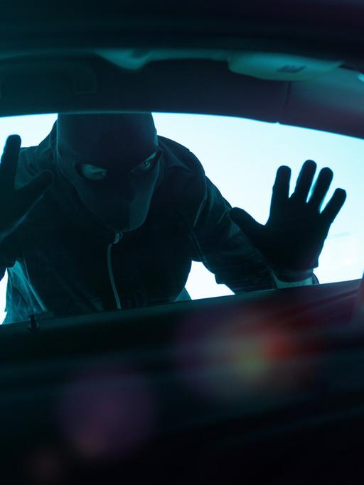 Ein Mann mit schwarzer Maske und Handschuhen steht bedrohlich an einem Autofenster.