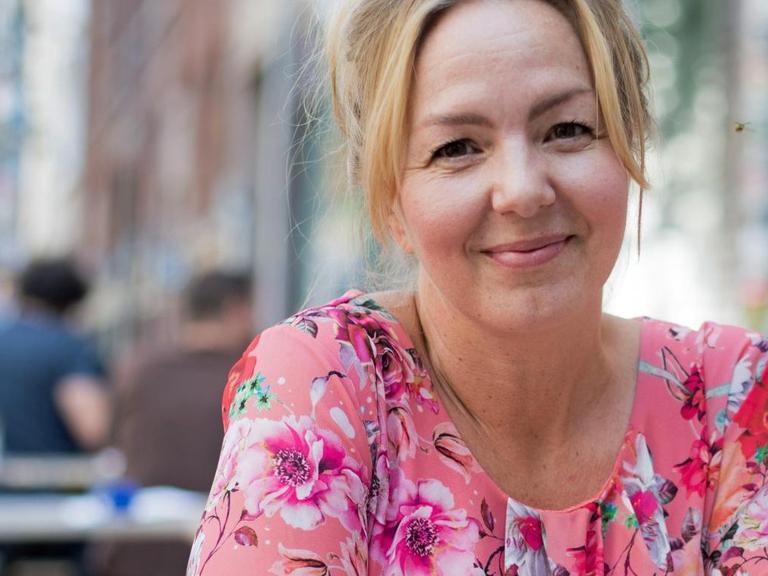 Die Schriftstellerin Simone Buchholz sitzt in blumiger Bluse an einem Tisch vor einem Straßencafé und lächelt in die Kamera.