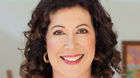 Eine Frau lächelt auf einer Porträtaufnahme frontal in die Kamera. Sie hat dunkelbraune Locken, trägt rot-pinken Lippenstift und goldene Ohrringe.