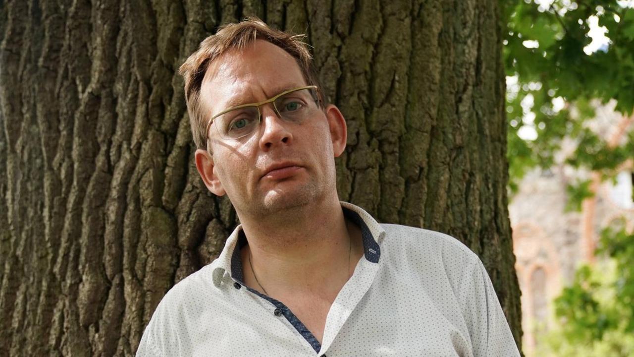 Clemens Meyer porträtiert am Rande einer Lesung auf dem Gut Bülowsiege im August 2019. Er steht im Halbschatten vor einem Baumstamm mit strengem Blick.