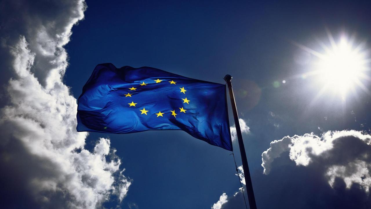 Die Fahne der Europäischen Union weht im Wind und wird von der Sonne angestrahlt. Gewitterwolken ziehen vorbei.