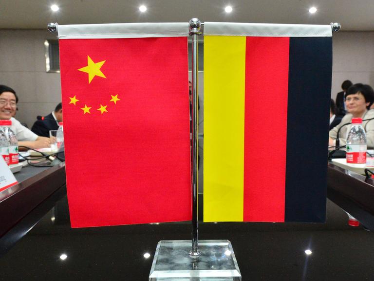 Eine Miniatur-Chinaflagge hängt neben einer Miniatur-Deutschland-Flagge an einem Ständer, der auf einem Tisch steht. Im Hintergrund sitzen Menschen am Tisch.