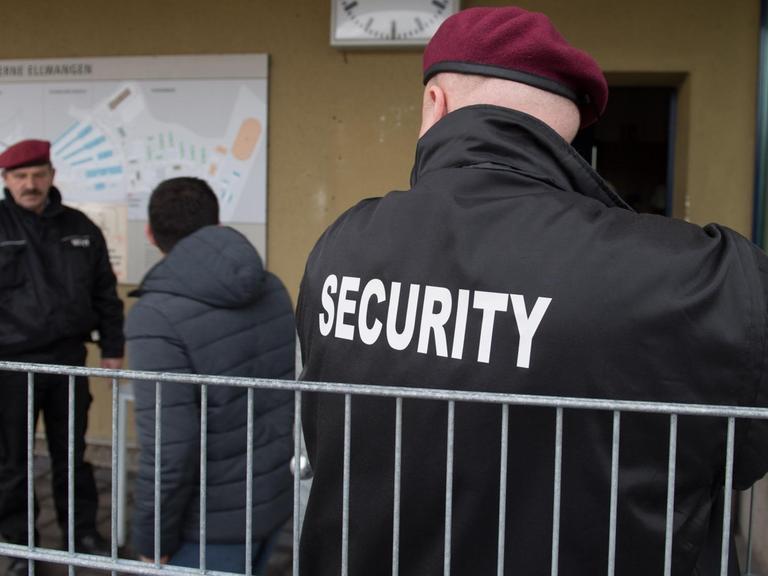 Zwei Sicherheitskräfte des Sicherheitsdienstleisters Siba Security GmbH, stehen im Februar in der Landeserstaufnahmeeinrichtung (LEA) in der Reinhardt-Kaserne in Ellwangen (Baden-Württemberg) an der Pforte, in der Mitte geht ein Flüchtling vorbei.