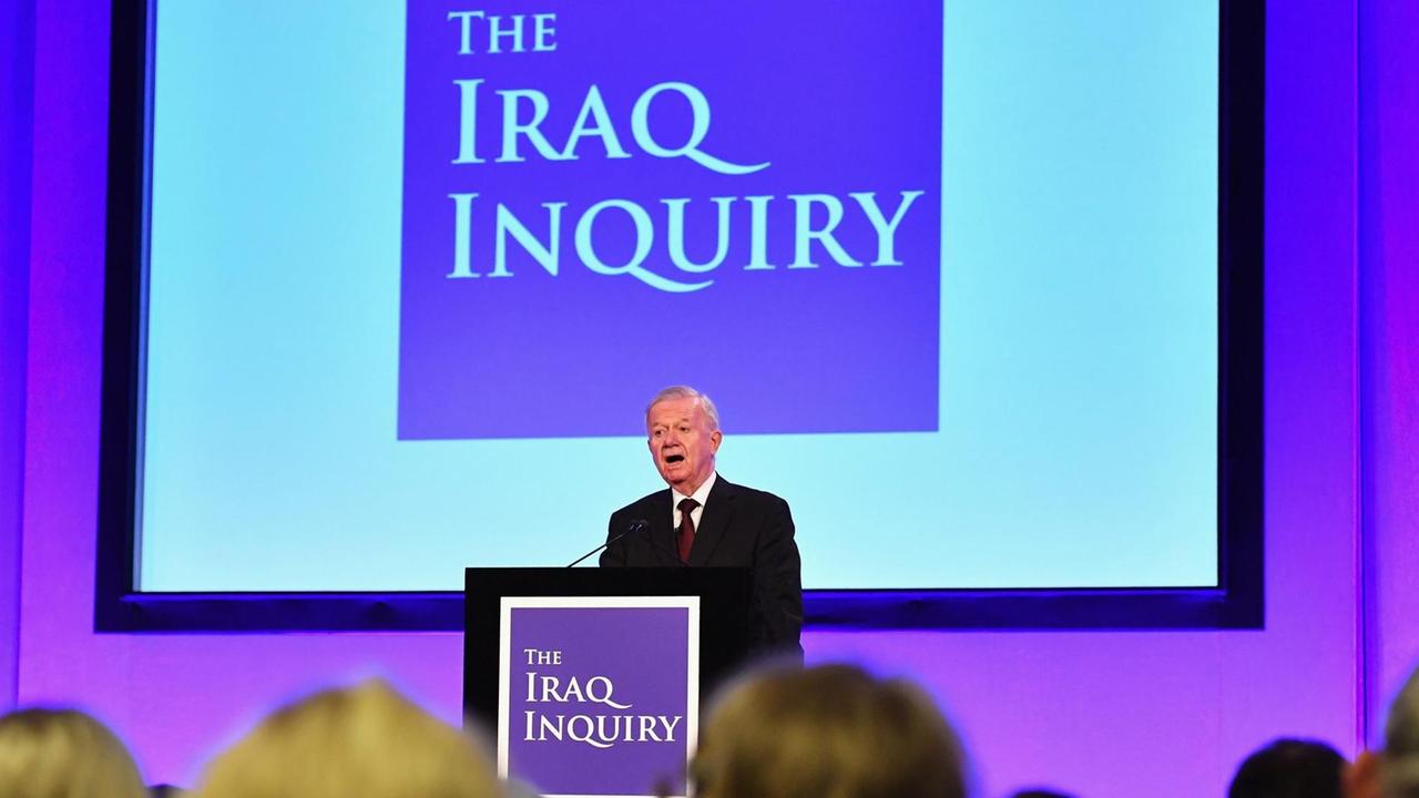 Chilcot steht an einem Rednerpult vor einer lila angestrahlten Wand mit einer großen Videotafel. Darauf steht "The Iraq Inquiry".