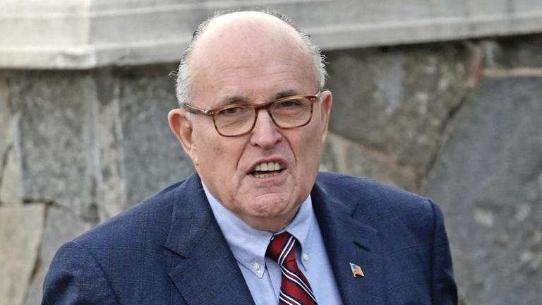 Rudy Giuliani trägt ein blaues Sakko, ein hellblaues Hemd und eine rot gemusterte Krawatte. Er schaut in die Kamera und steht vor einer Mauer.