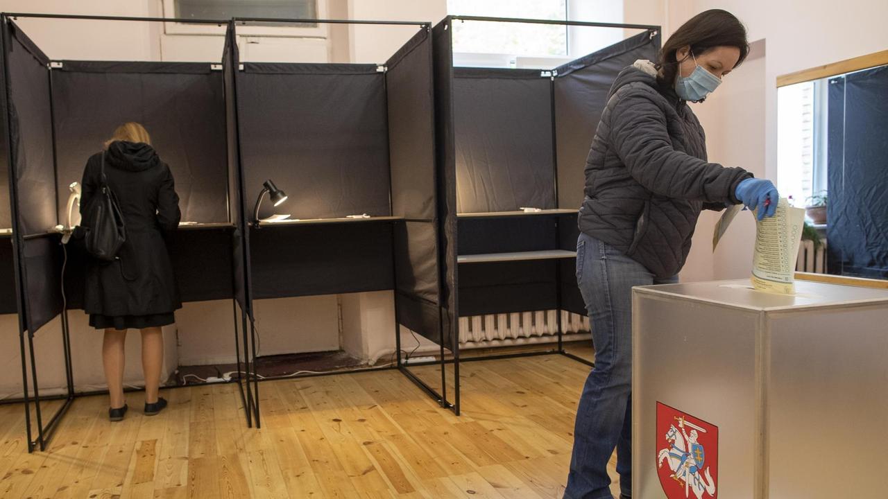 Die Frau wirft den Zettel in die milchig-transparente Wahlurne mit dem Wappen Litauens. Hinter ihr drei Wahlkabinen, in denen eine weitere Frau gerade ihren Stimmzettel ausfüllt. 