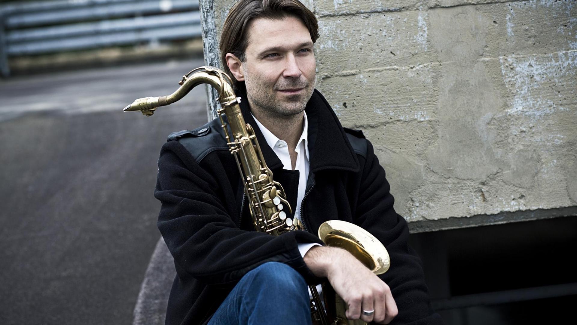 Der Jazzkomponist Heiner Schmitz sitzt mit einem Saxophon an eine Mauer gelehnt