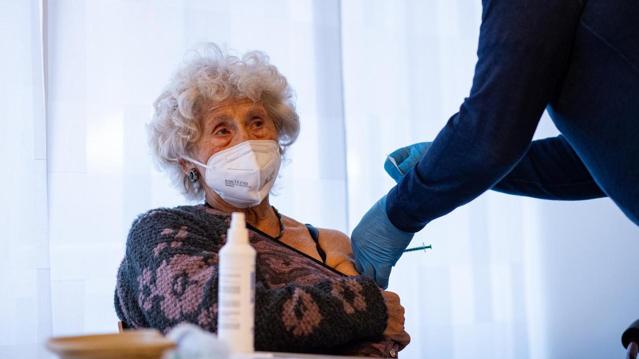 Eine ältere Dame mit grauen Locken und weißem Mundschutz hat den Ausschnitt ihres Pullovers bis über den linken Oberarm gezogen, so dass ein Arzt sie dort impfen kann.