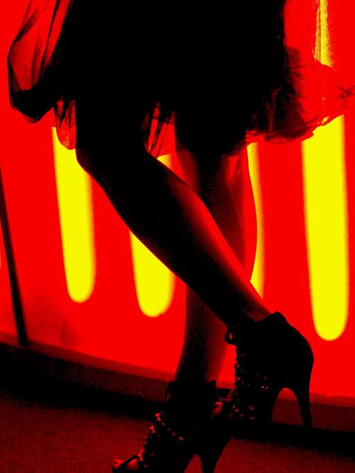 Eine Frau mit einem teilweise transparenten Rock steht mit High-Heels an einer rot beleuchteten Bar und unterhält sich mit einem Mann.
