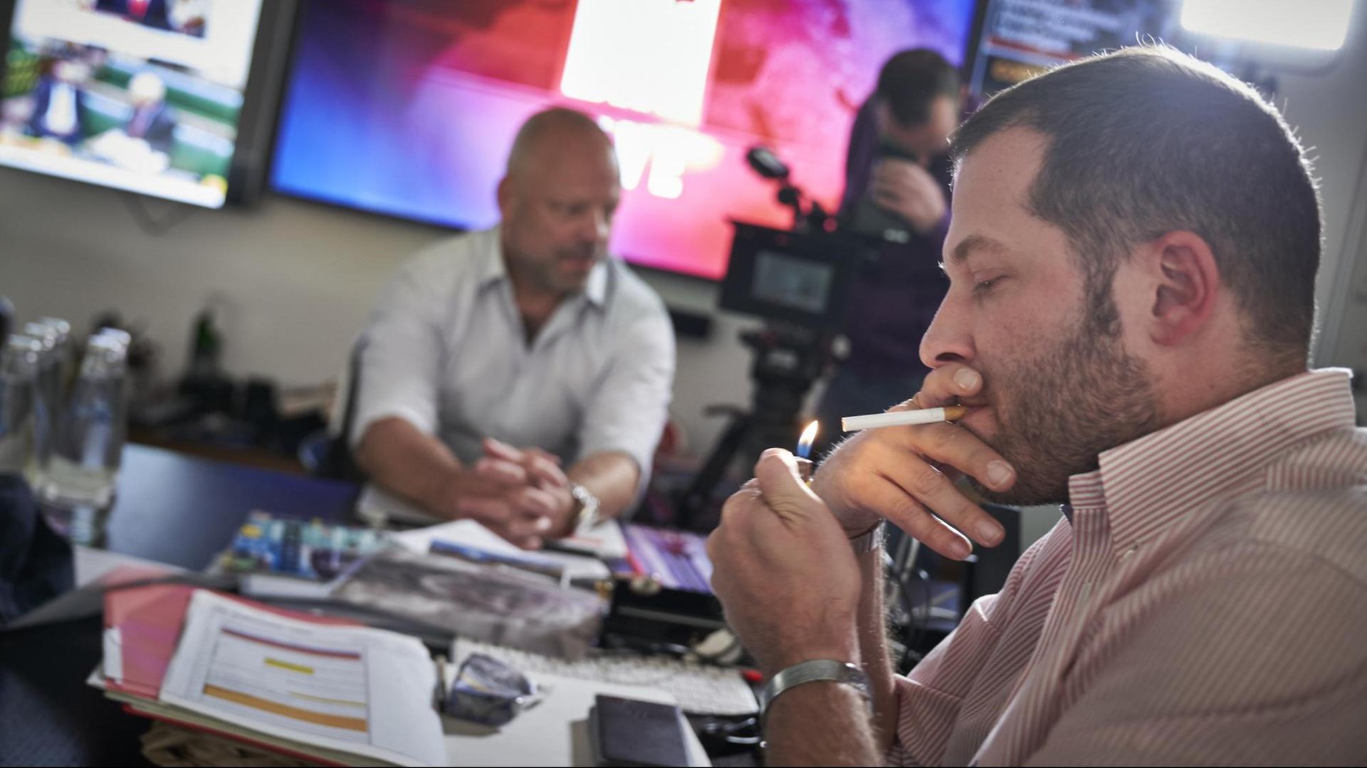 Julian Reichelt, Chefredakteur der Bild-Zeitung, zündet sich eine Zigarette an. Im Hintergrund sitzt ein anderen Mann an einem Tisch, ein weiterer Mann hantiert mit einer Kamera.