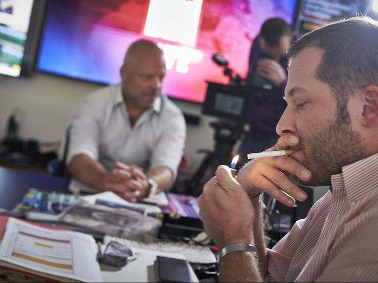 Julian Reichelt, Chefredakteur der Bild-Zeitung, zündet sich eine Zigarette an. Im Hintergrund sitzt ein anderen Mann an einem Tisch, ein weiterer Mann hantiert mit einer Kamera.