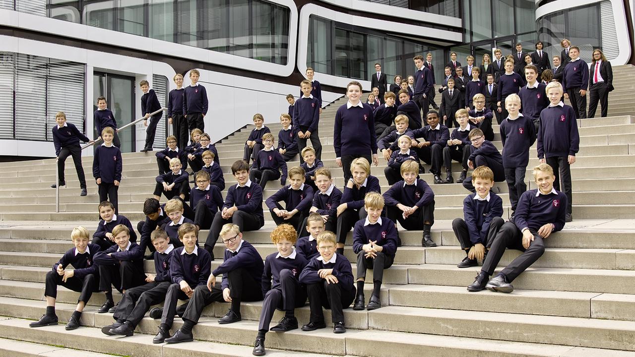 Viele Kinder unterschiedlicher Altersgruppen sitzen in Chorkleidung auf den Stufen ihres Schulgebäudes.