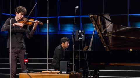 Ein junger Geiger und ein junger Pianist musizieren vor Mikrofonen und einem rot leuchtenden Aufnahmelicht in einem holzgetäfelten Saal.