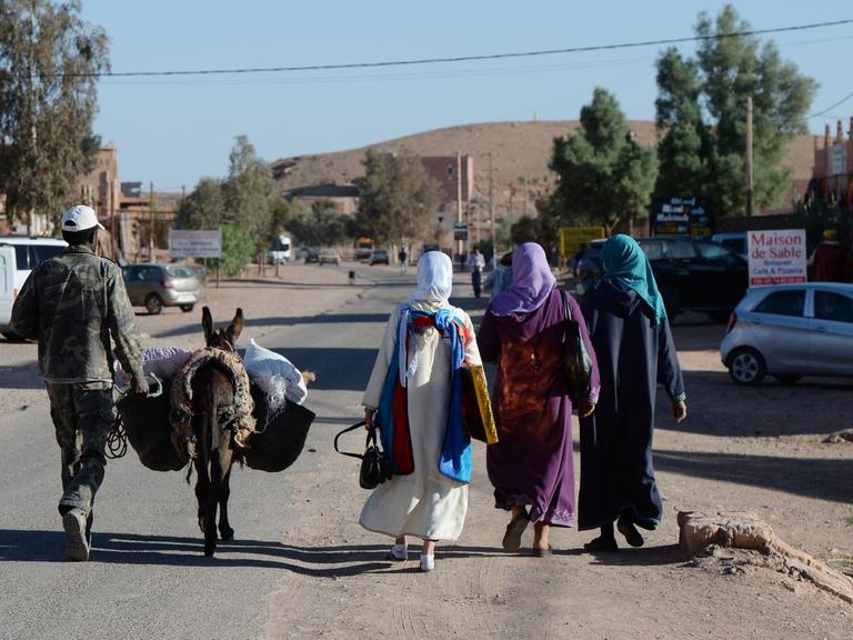Ein Mann mit einem Esel und drei Frauen in traditioneller Kleidung gehen auf einer Straße bei Ait-Ben-Haddou (Marokko).