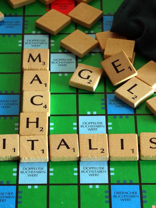 Auf einem Scrabble-Brett sind mit Buchstaben-Steinen die Begriffe "Kapitalismus" und "Macht" zusammengefügt, scheinbar wahllos im Hintergrund liegende Steine bilden das Wort "Geld".