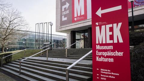 Treppenaufgang zum Eingang des Museums für Europäische Kulturen. Vorne rechts steht eine rotes Schild mit der Aufschrift "MEK Museum Europöischer Kulturen", dahinter ist ein Teil des Gebäudes zu sehen.