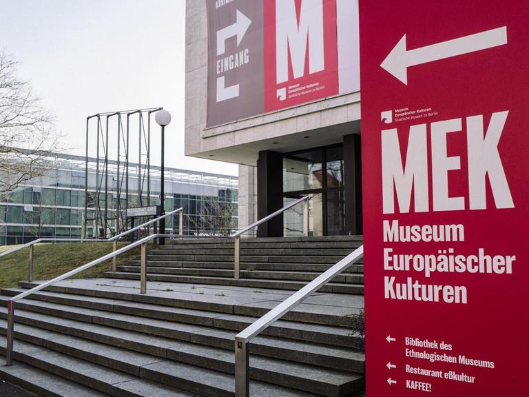 Treppenaufgang zum Eingang des Museums für Europäische Kulturen. Vorne rechts steht eine rotes Schild mit der Aufschrift "MEK Museum Europöischer Kulturen", dahinter ist ein Teil des Gebäudes zu sehen.