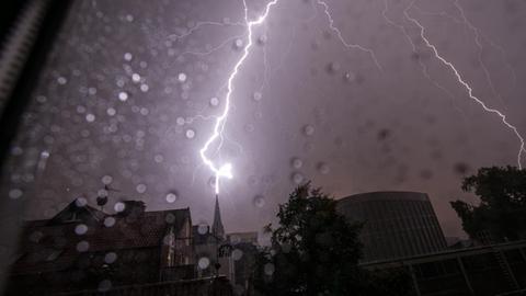 Blick durch ein regennasses Fenster auf einen Kirchturm, in den gerade der Blitz einschlägt.