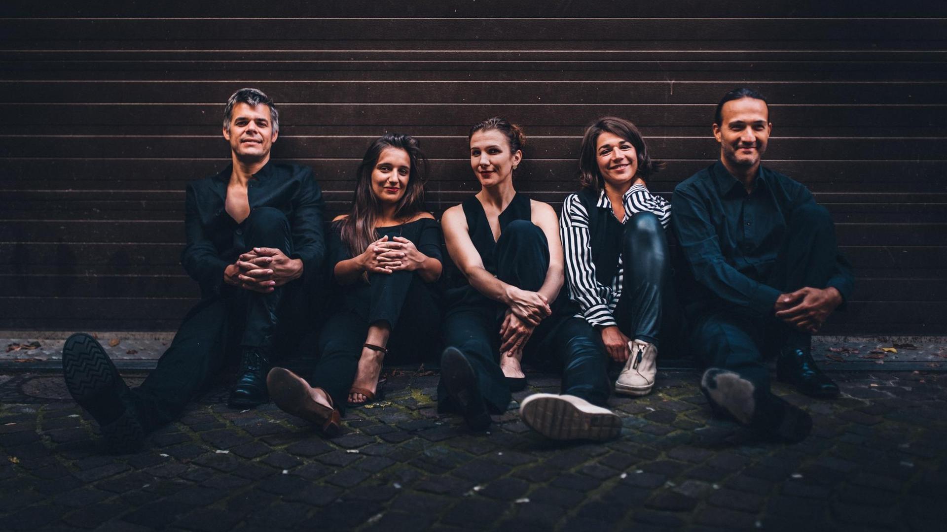 Sieben Musikerinnen und Musiker in legerer Kleidung sitzen nebeneinander vor einer Wand, sie haben alle fröhliche Gesichter.